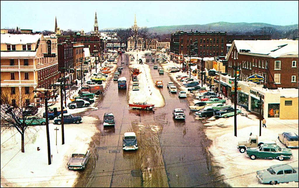 A colored photograph of a wide street bordered by several multi-storied buildings. All of the buildings and sidewalks are covered in snow. Cars are parked on both sides of the street. There are also some cars driving on both sides of the road. 