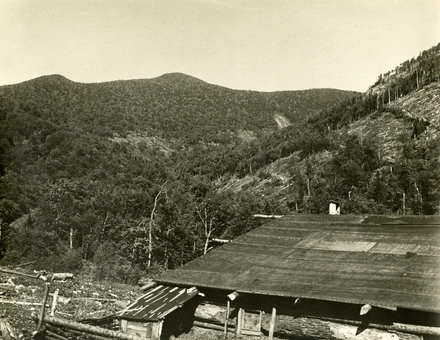 A black and white photograph shows a view of a mountain in the  background with a small building surrounded by the some cleared land in the foreground. To the right, a partially cleared mountainside is visible. 