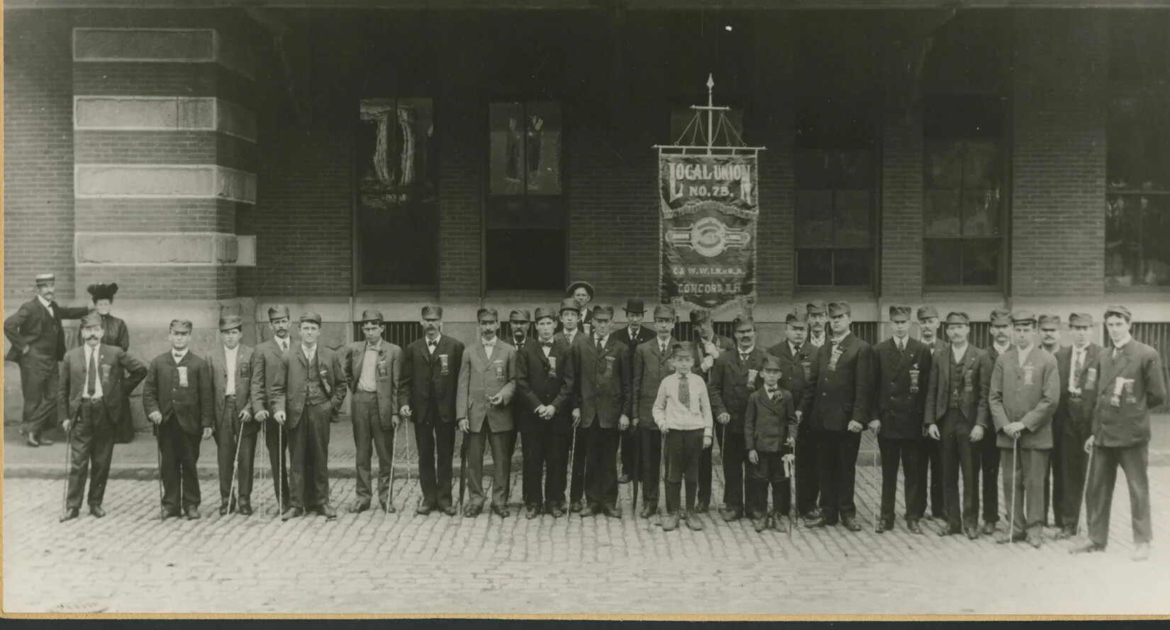 A black and white photograph of a group of men and two young boys posed in a line, starring at the camera. All of the men wear ribbons on the fronts of their suit jackets. Someone holds up a large sign in the back of the group, which reads "LOCAL UNION/NO. 75/CONCORD, N.H."