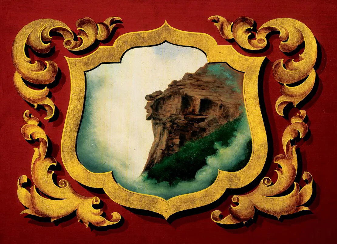 An ornate painting shows a picture framed by gold decorations with red background. The picture shows the side of a mountain, and the upper third of the rocky cliff appears to look like the profile of a man. 