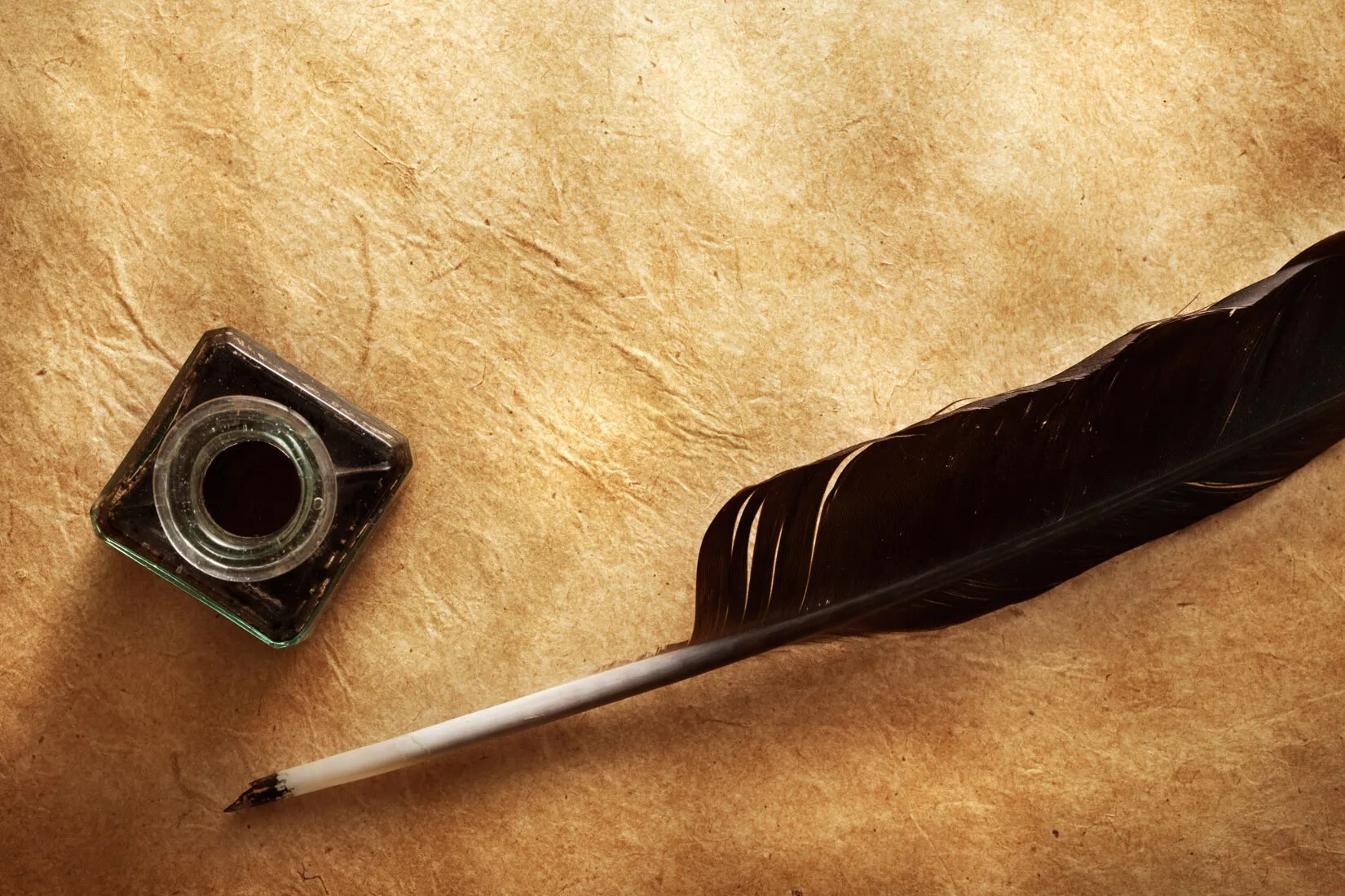 A brown quill and inkwell lie on top of a blank document made from browning paper.