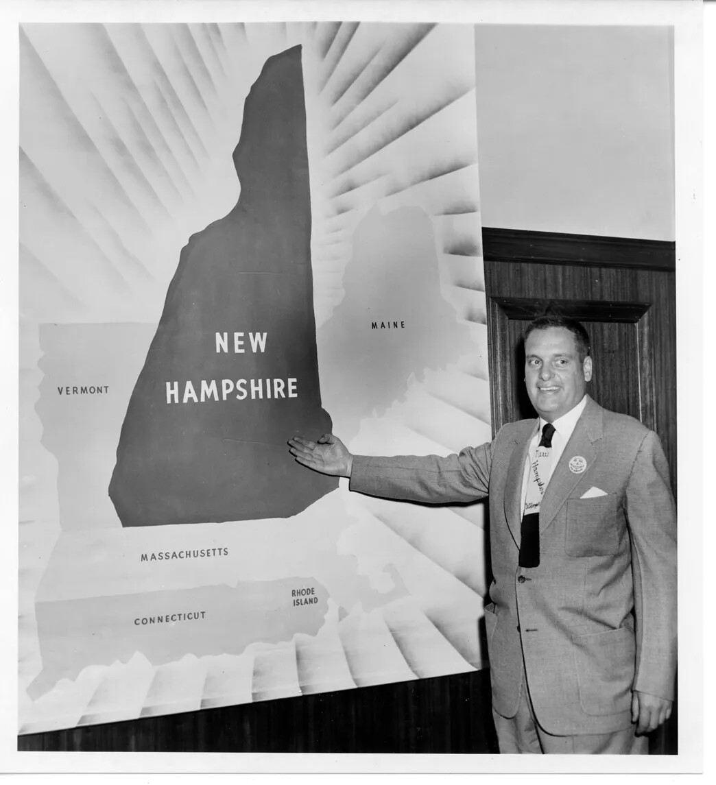 In a photograph, a man stands in a suit and gestures to a map behind him. The map shows New England, and the state of New Hampshire is larger than usual.