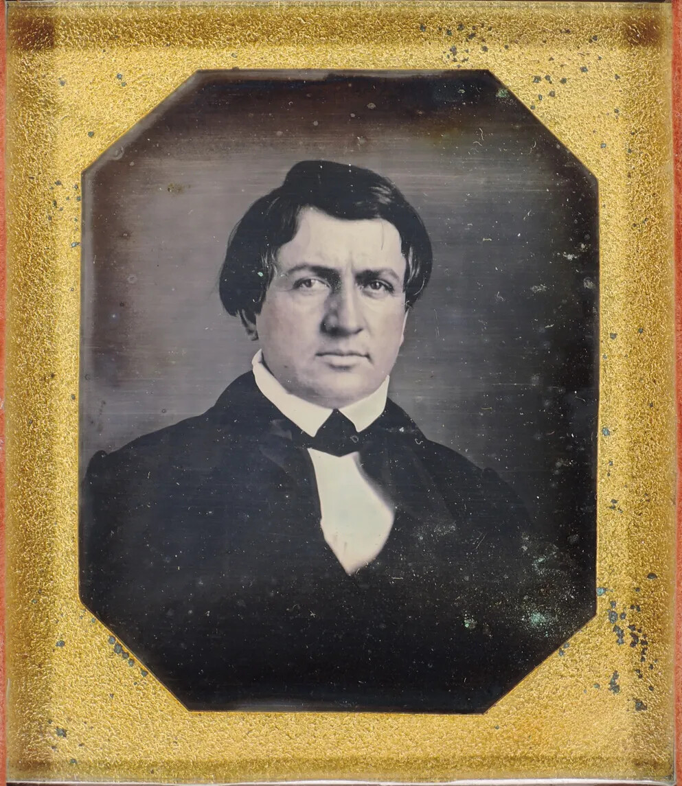 A black and white photograph of a man looking at the viewer. He wears a white collared shirt and a black necktie and jacket.