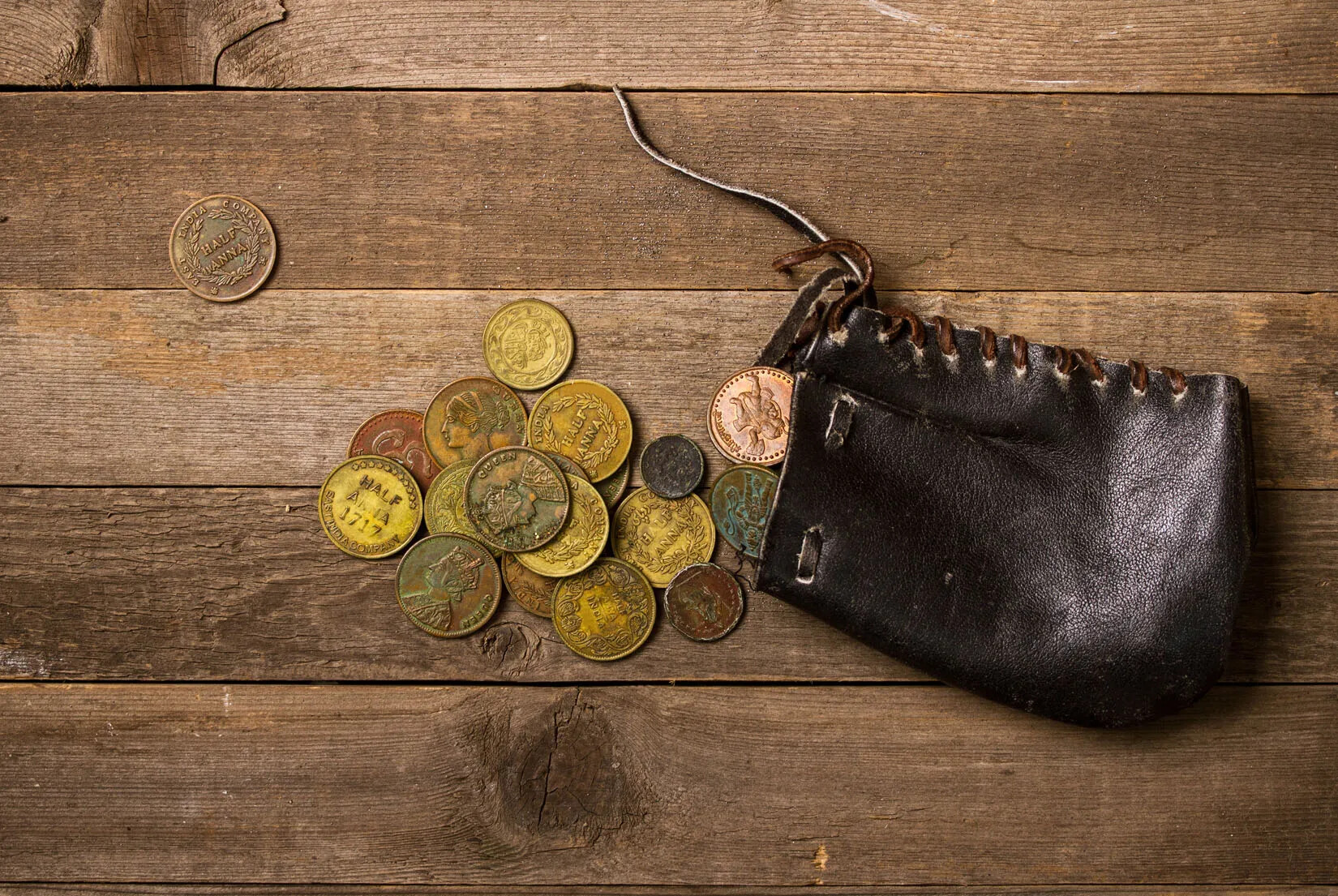 A leather pouch lying on a wooden table, laced up by leather ties. Pouring out of the pouch are several gold and brass colored coins.