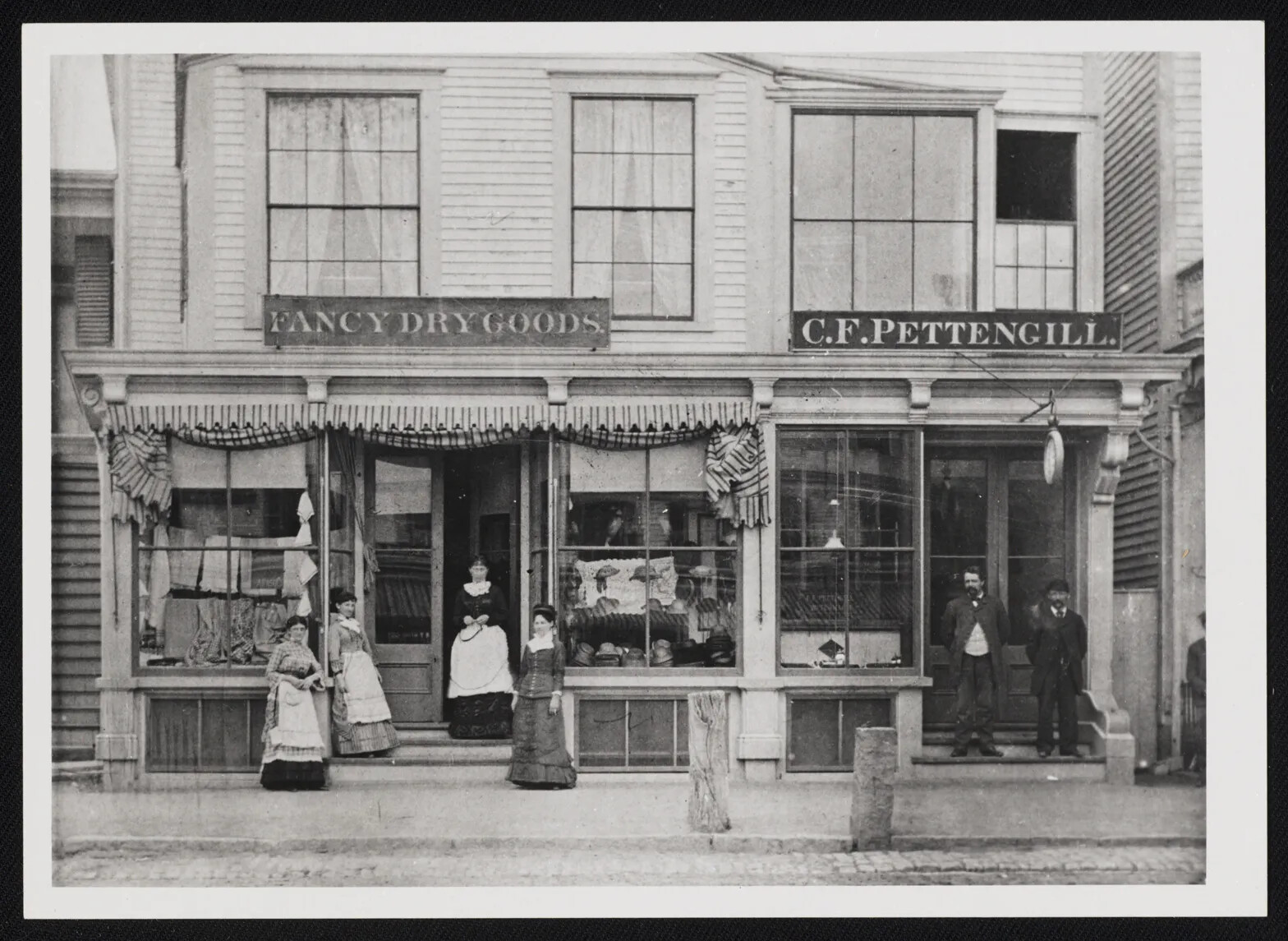 A black and white photograph of the exterior of a store. The store is two-stories tall and has two entrances. Two men stand in front of the entrance to the right, underneath a sign that reads "C.F. PETTENGILL." and four women stand in front of the entrance to the left, underneath a sign that reads "FANCY DRY GOODS." The women wear long dresses with long sleeves and the men wear jackets and pants.