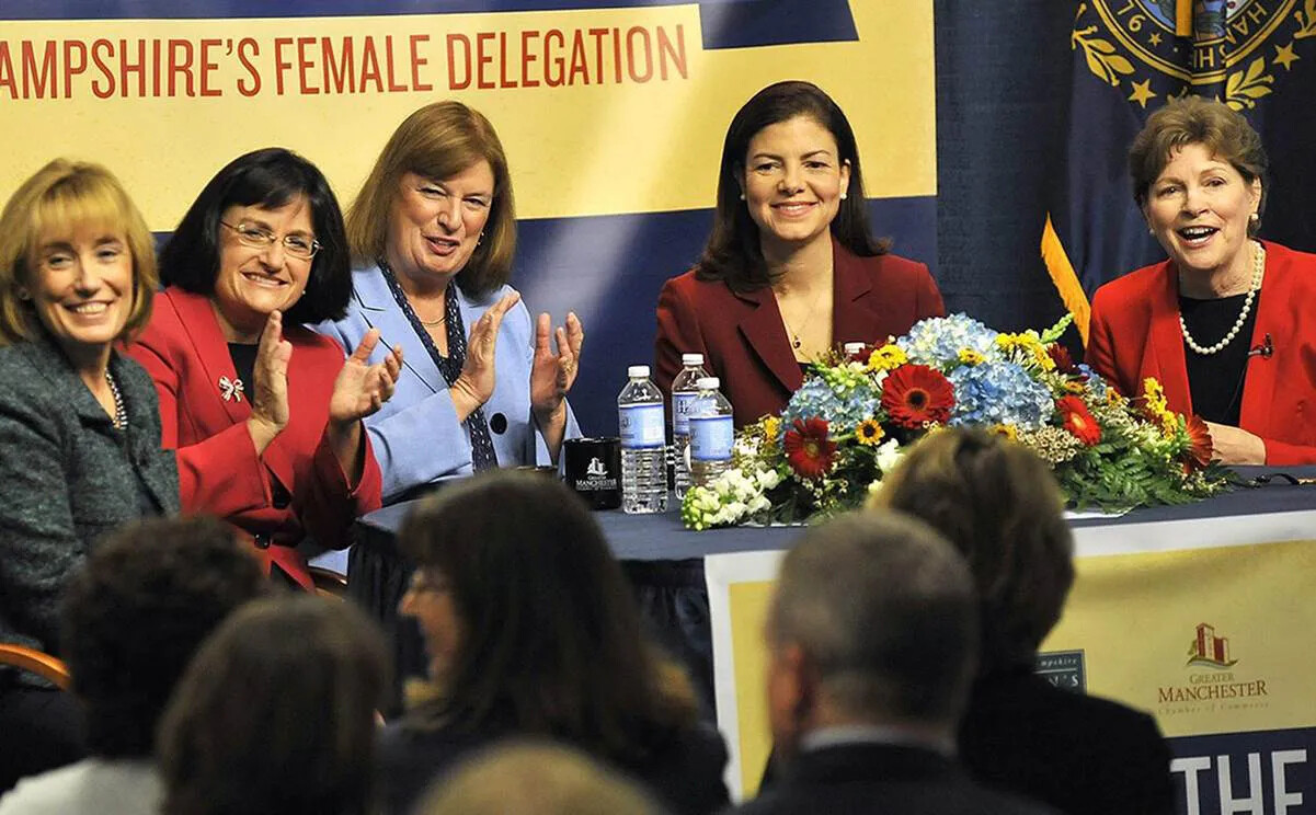 A colored photograph of five white women seated behind a table. They are all facing an audience and are smiling and clapping their hands.