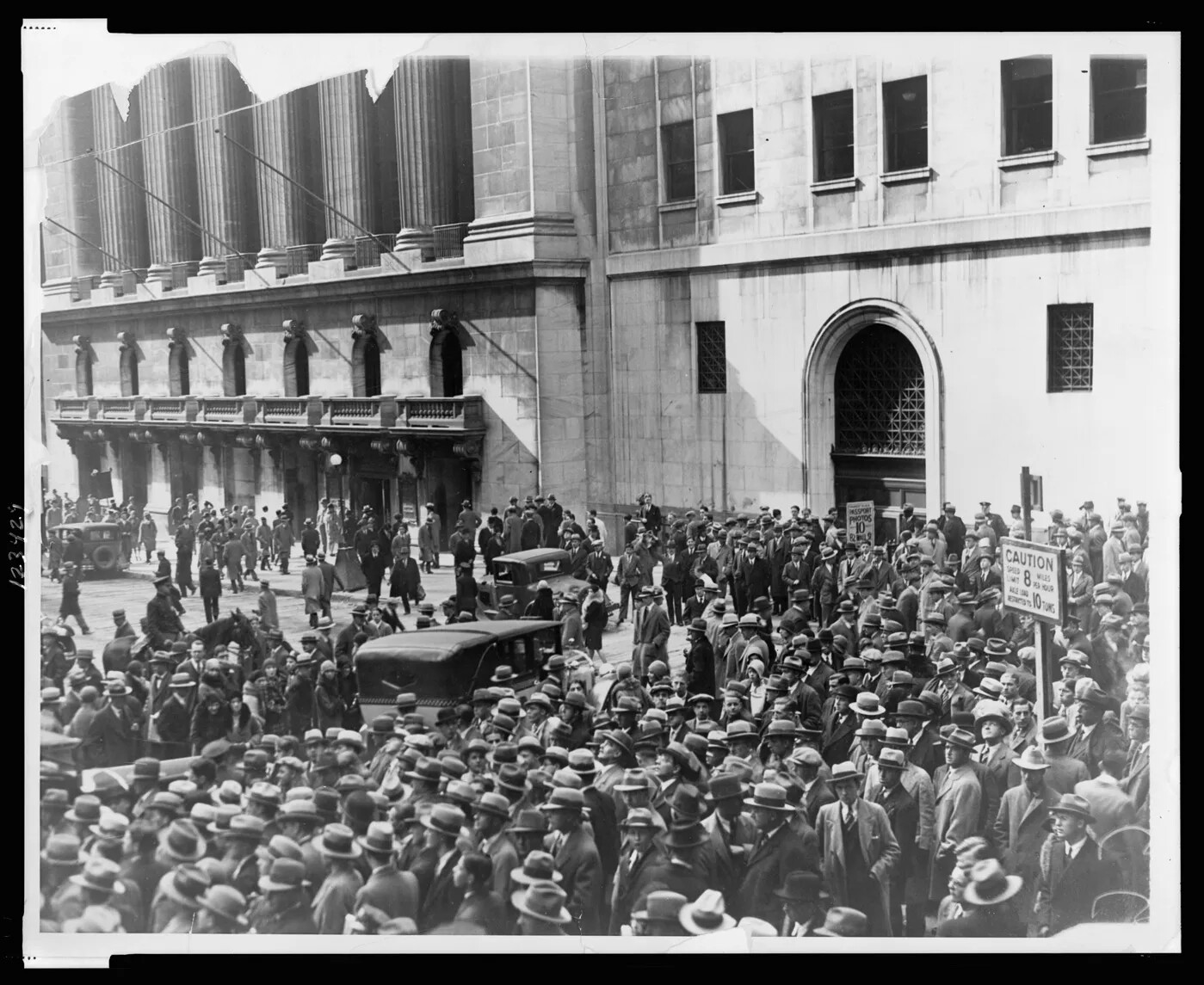 Part of a large stately building with columns and multiple entrances is seen in a black and white photograph. A large crowd of people have gathered on the street, and most wear wide brimmed hats, suits, and long jackets. Three 1930s cars are among the crowd. 