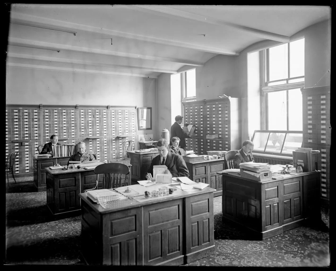 A black and white photograph of six men working in an office. Five of the men sit at large desks and one man stands in the back of the room and looks through files at a cabinet. The room is lined with several tall cabinets with multiple drawers.