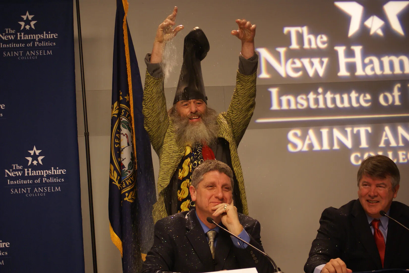 A photograph of a man with a boot on his head, an animal-print jacket, three ties, and long facial hair throws glitter over the head of a seated man. Two seated men in ties and jackets have a small grin on their faces.  