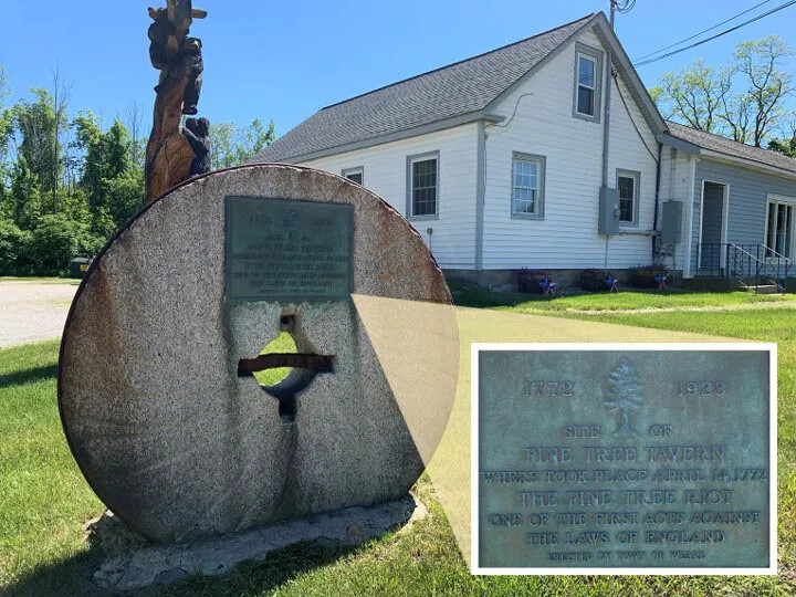 A large stone wheel stands upright and has a hole in the middle. On the upper rim of the wheel, a large plaque is placed above the hole. The words on the plaque are raised engravings. The words read: "1772 1928/SITE OF/PINE TREE TAVERN/WHERE TOOK PLACE APRIL 14, 1772/THE PINE TREE RIOT/ONE OF THE FIRST ACTS AGAINST/THE LAWS OF ENGLAND/ERECTED BY THE TOWN OF WEARE."