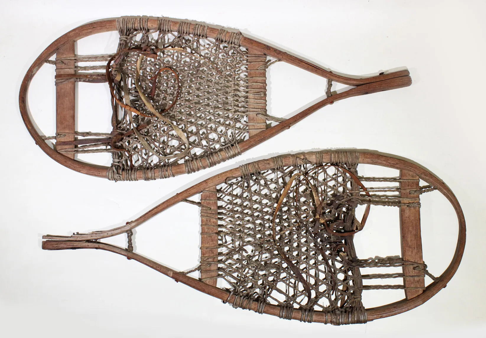 A pair of flat, wooden, tear-shaped snow-shoes. There is a woven netting of rope on the inside of the shoe, which is tied to two small wooden boards that lie vertically across the shoe.