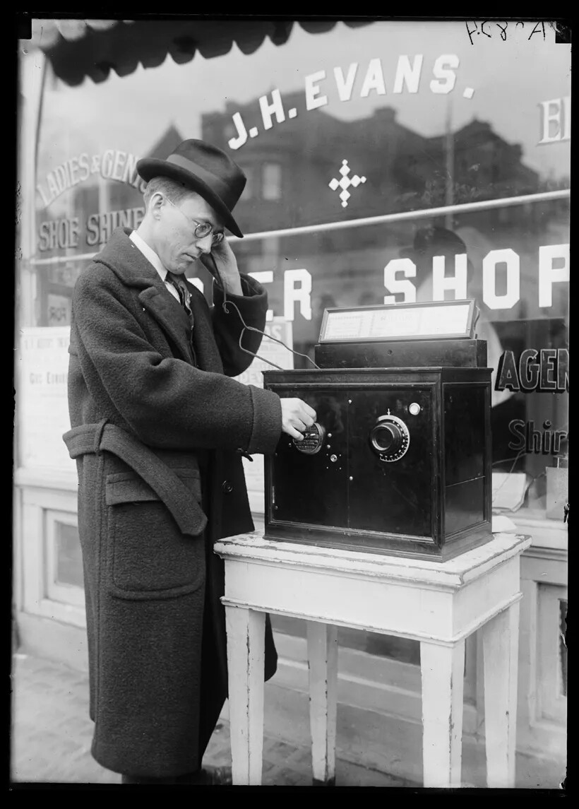 A black and white photograph shows a man in a long coat and hat standing outside a store on a street. Next to him is a table with a large black box on it. On the box are two circles with markings. He is holding something connected with a wire to the black box up to his ear, and touching one of the circles with the other hand. He is looking down at the box.. 