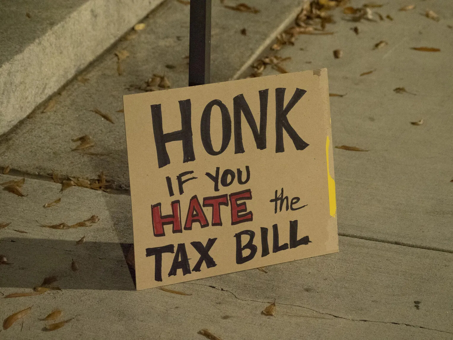 A colored photograph of a cardboard sign leaning up against a street sign on a sidewalk. The sign reads "HONK/IF YOU/HATE the/TAX BILL."