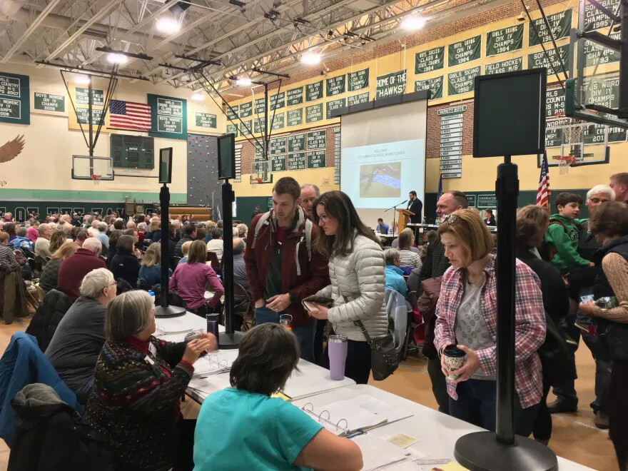 A colored photograph of a gymnasium filled with people sitting in rows. At the forefront of the photograph is a table, which has three people seated on one side and three people standing on the other.