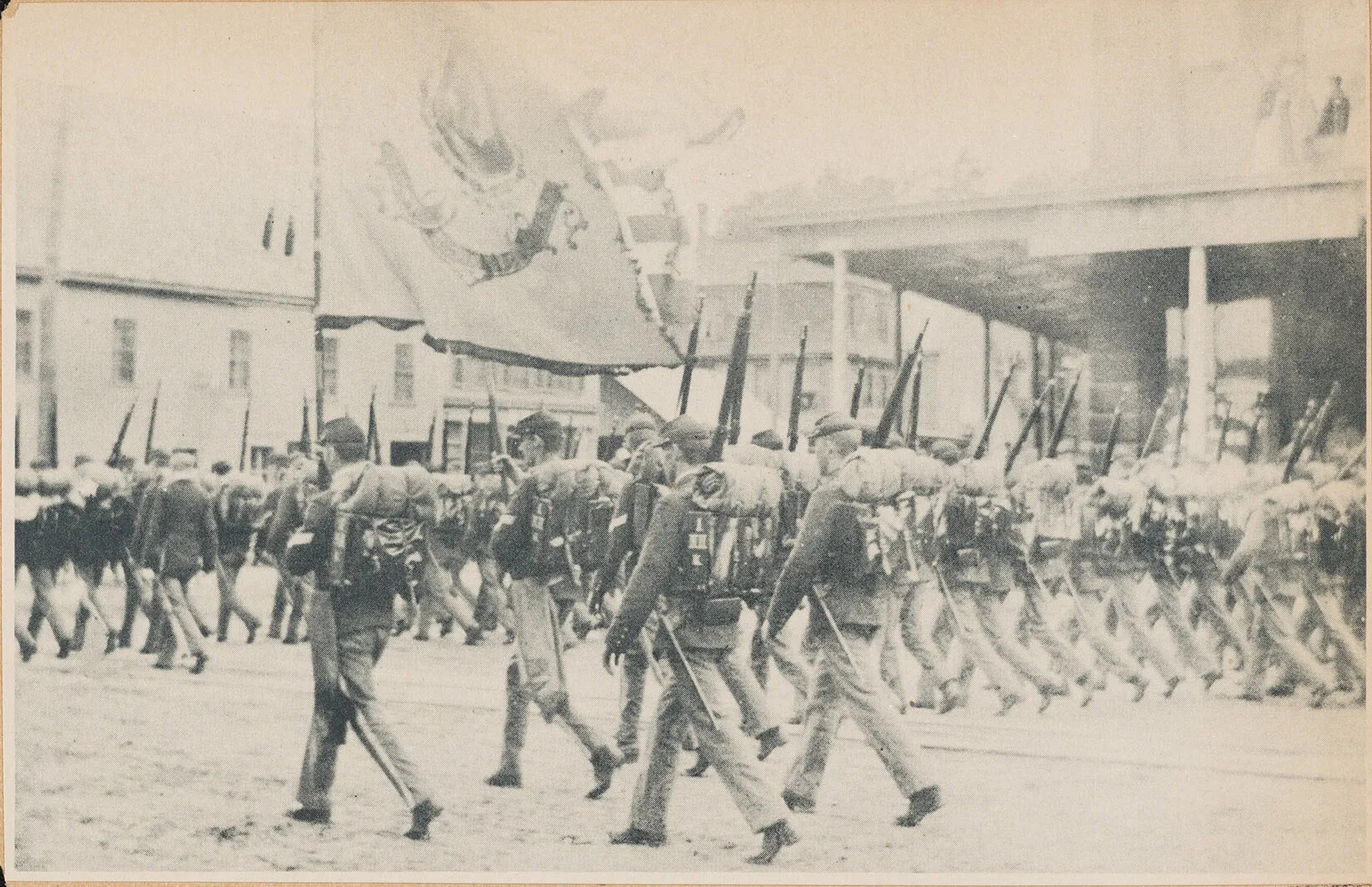 A black and white photograph of a group of men marching in straight lines. They all carry muskets and backpacks with rolled-up blankets.