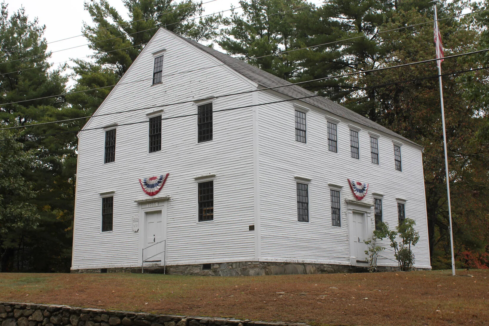 A colored photograph of a two-storied white building with a pitched roof and attic space. Semi-circular American flags hang over the front and side doors. A flag pole with an American flag stands to the building's right.