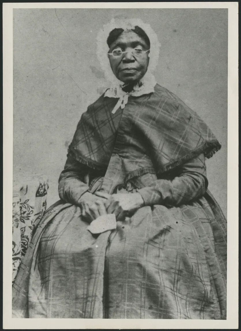 A black and white photograph of a Black woman sitting with her hands placed on her lap. She wears glasses, a long dress with long sleeves, a shoulder-length cloak, and a bonnet.