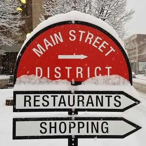 A colored photograph of a street sign. The top part of the sign is a red semi-circle and is labeled" "MAIN STREET/DISTRICT" in white lettering. The lower part of the sign are two white and black arrows, pointed to the right, which are labeled: "RESTAURANTS" and "SHOPPING."