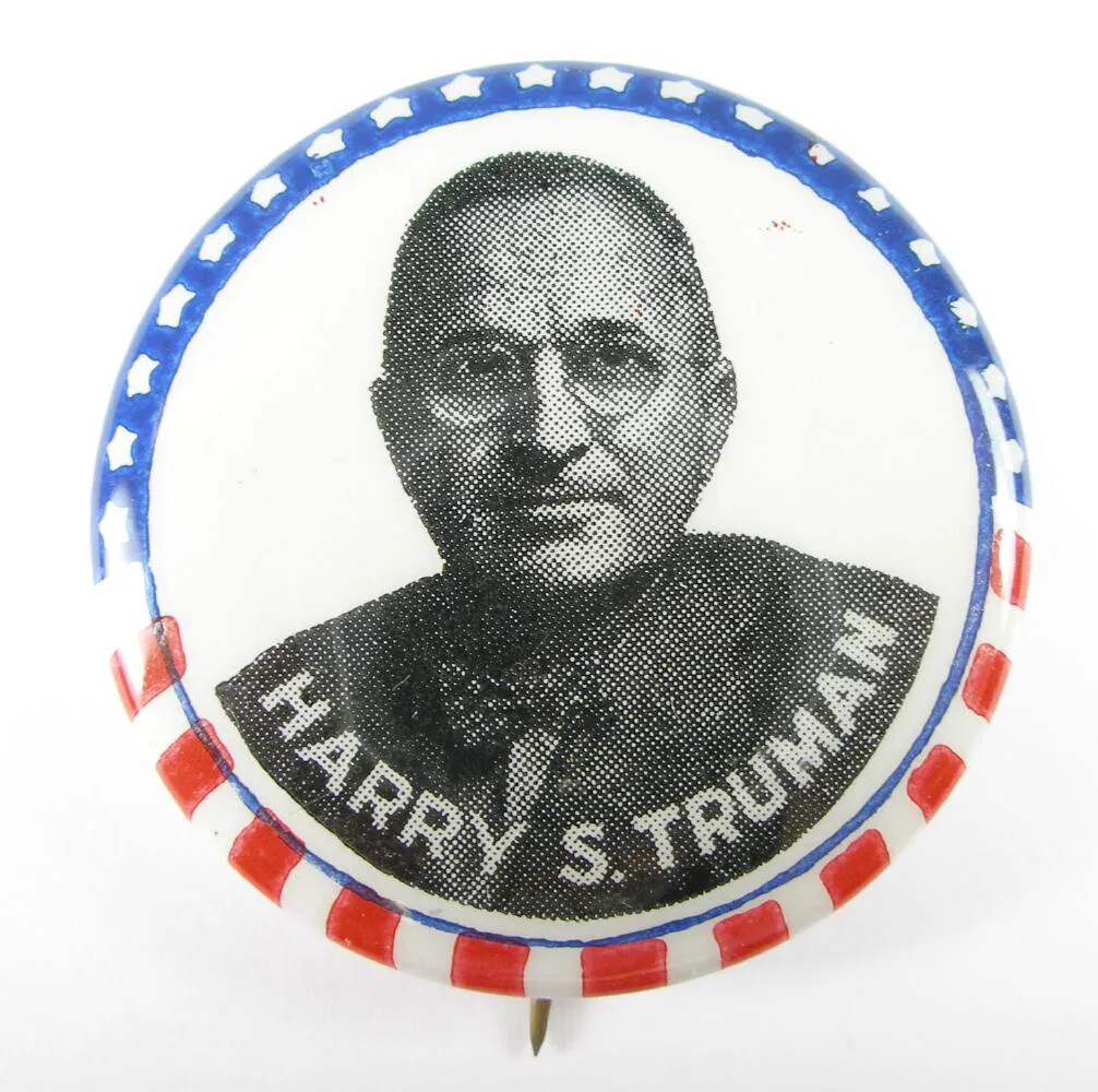 A circular button with a man facing forward. He is wearing round glasses, a suit jacket, and a bow tie. Along the upper rim of the button is a blue line with white stars and along the bottom rim are alternating red and white  vertical stripes. The words "HARRY S. TRUMAN" are placed along the bottom rim and curve along with the edges of the button.