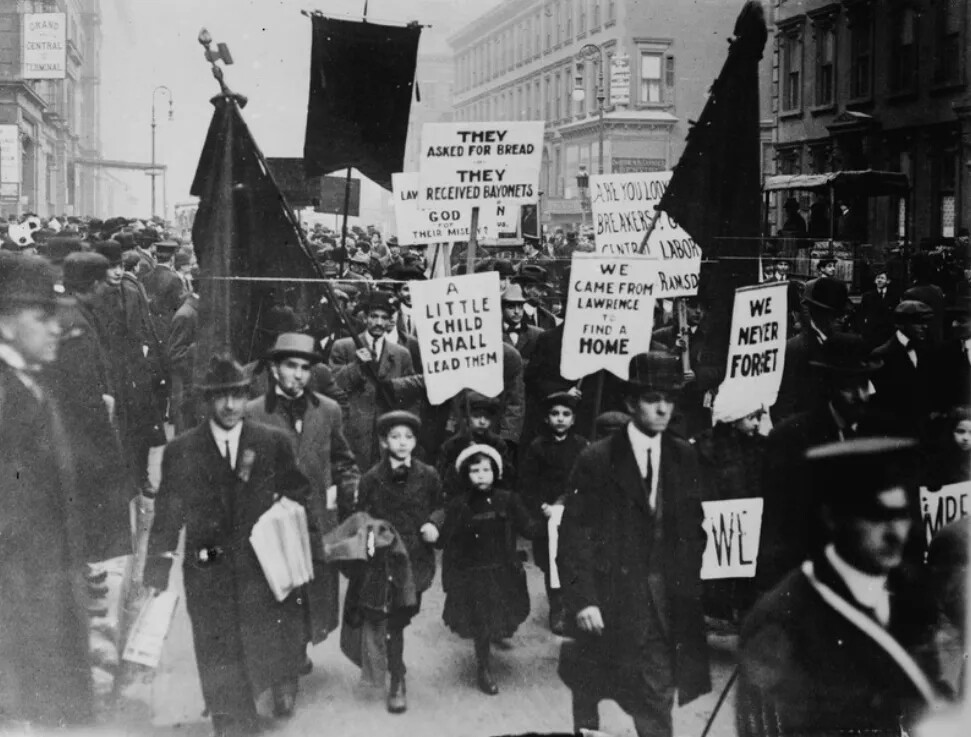 A black and white photograph of a group of men and children marching down a street. Many people carry signs on sticks and large, dark-colored flags.