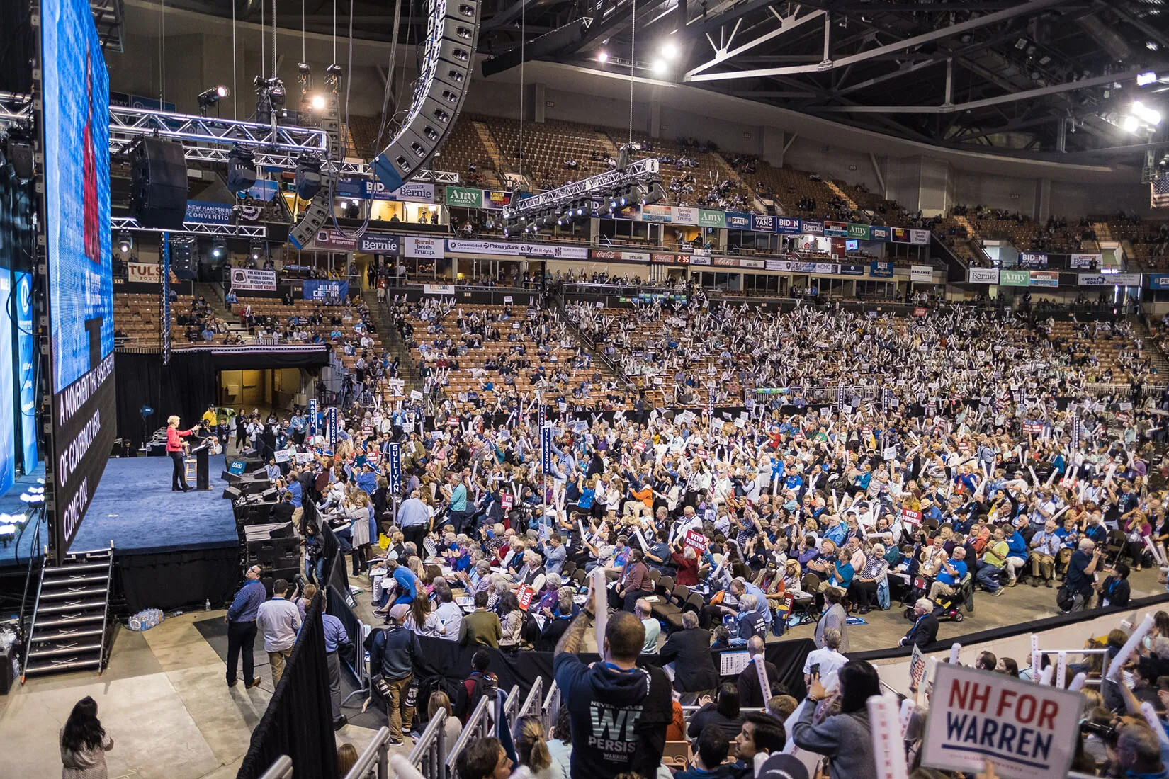 A colored photograph of an indoor stadium filled with people. They all face a woman who stands behind a podium on a stage.
