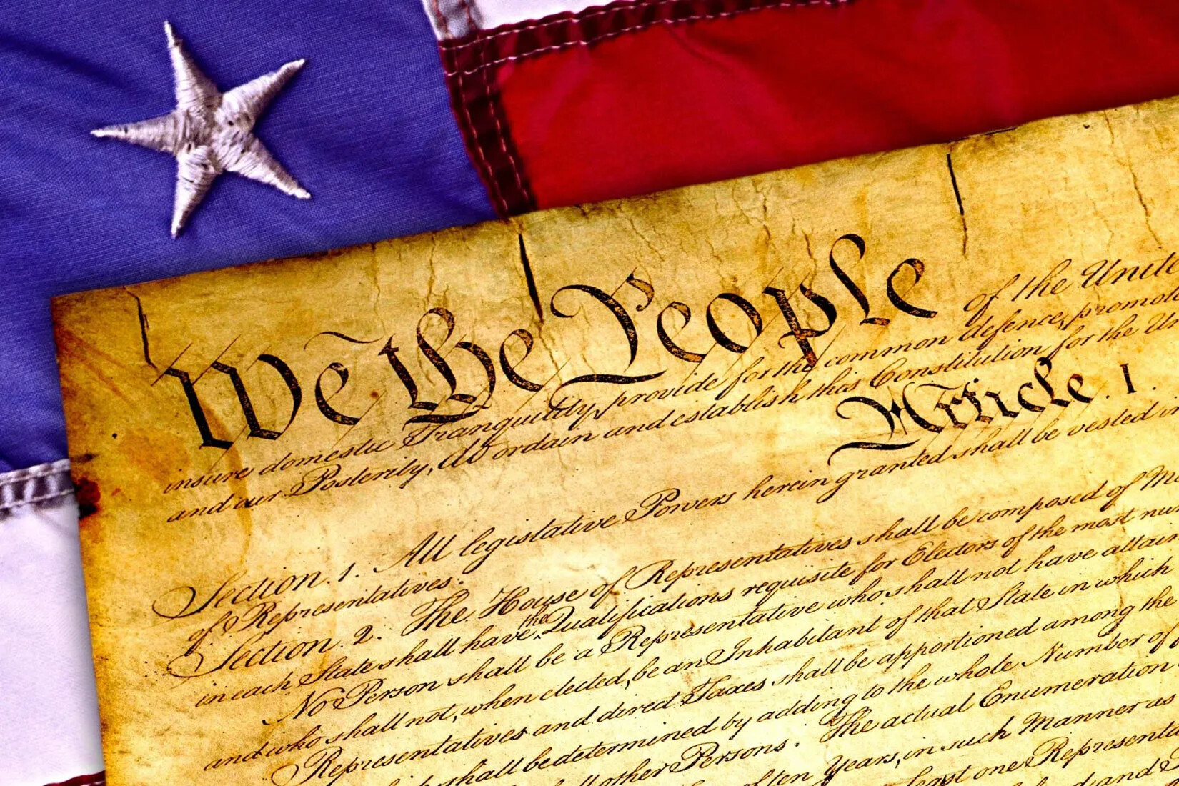A colored photograph of the corner an old document with the words "We the People" written in black ink in cursive handwritting. The document lies on top of a cropped American flag.