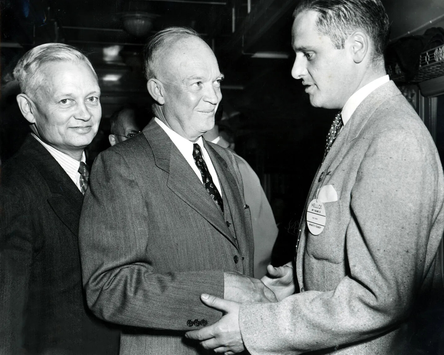 A black and white photograph of three white men in collared shirts, ties, and suit jackets. The man on the left looks directly at the camera, while the other two men shake hands and look at each other.
