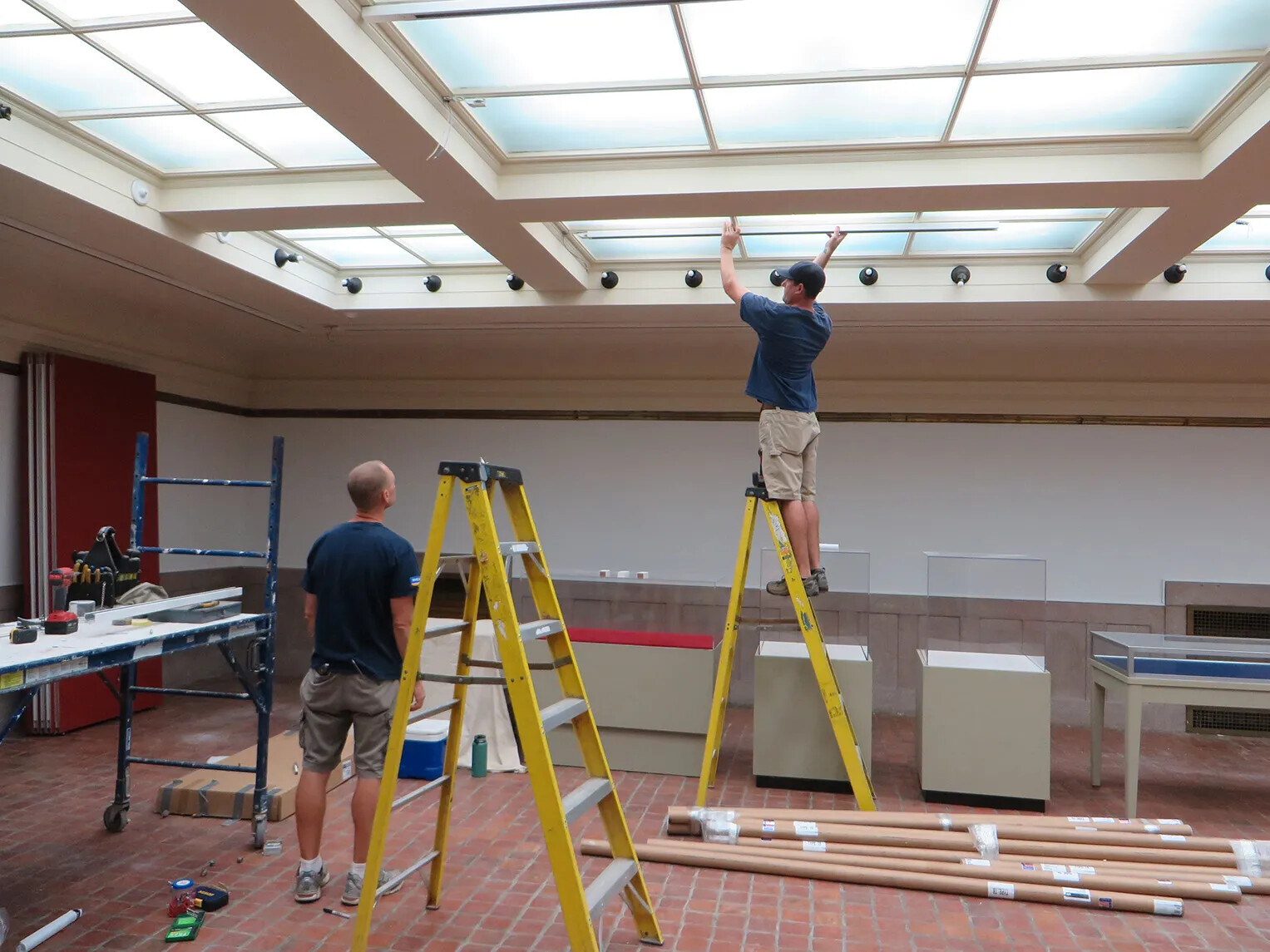 A colored photograph of two men in a room with a sky-light. One of the men stands on a yellow ladder and holds up a horiztonal piece of wood to the ceiling. The other man watches him from the ground.