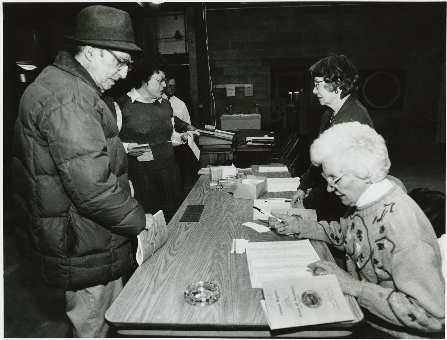 A black and white photograph of a group of people standing around a long table. At the front of the photograph, a man in a winter coat and a fedora stands on one side of the table and talks to a seated woman on the other side of the table, who is looking through a multi-paged document. In the background of the photograph, two other women stand on opposite sides of the table.