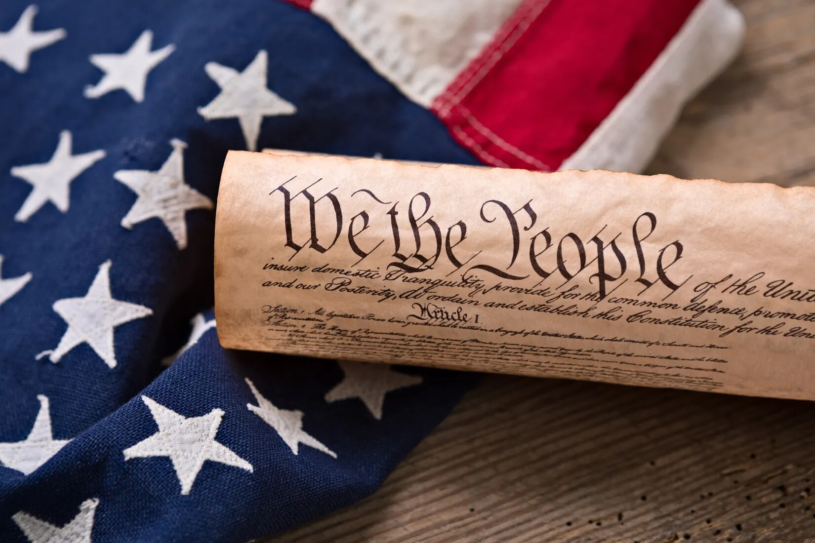 A colored photograph of the rolled up old document with the words "We the People" written in black ink in cursive handwritting. The document lies on top of a cropped American flag.