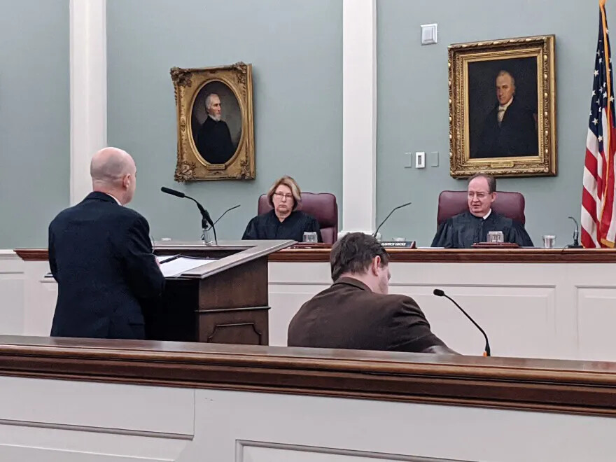 A colored photograph of a white man and a white woman in black robes sitting behind a high counter, facing the viewer. They look at a bald white man, who stands with his back to the viewer in front of a podium, and a seated man with brown hair, who also faces away from the viewer.