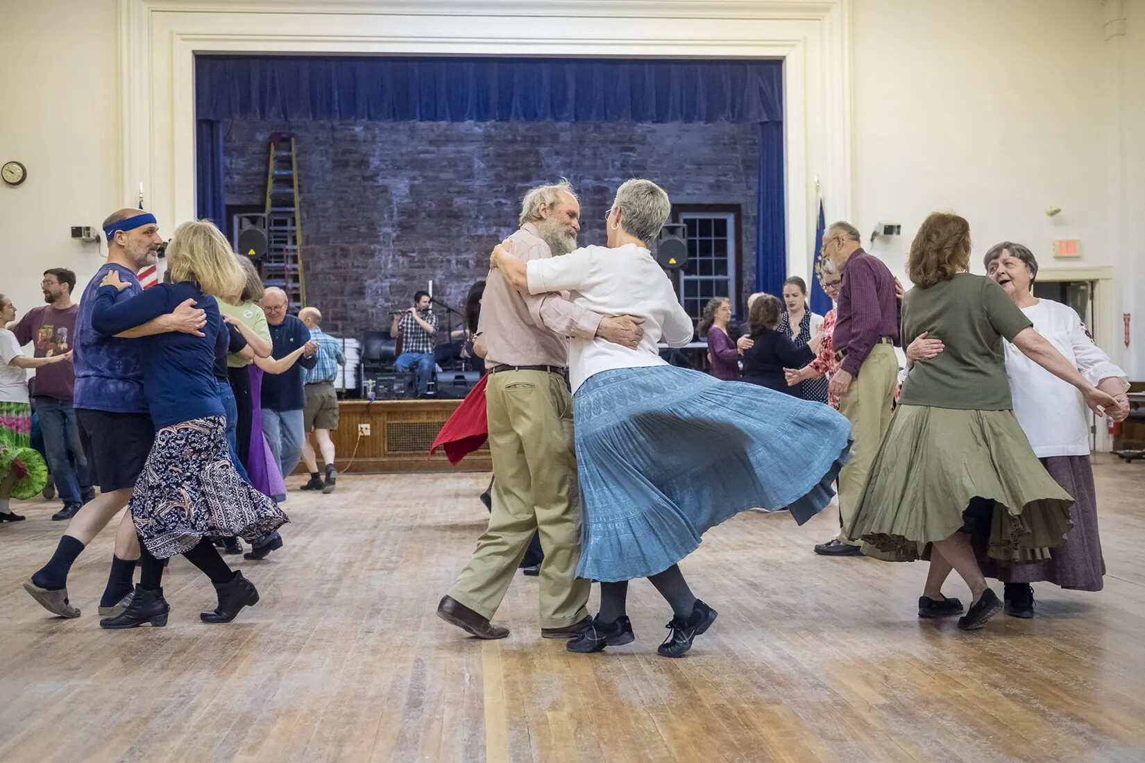A colored photograph of men and women dancing in pairs in a large room. At the back of the room is a stage. A musician playing the flute can be seen seated on the stage.