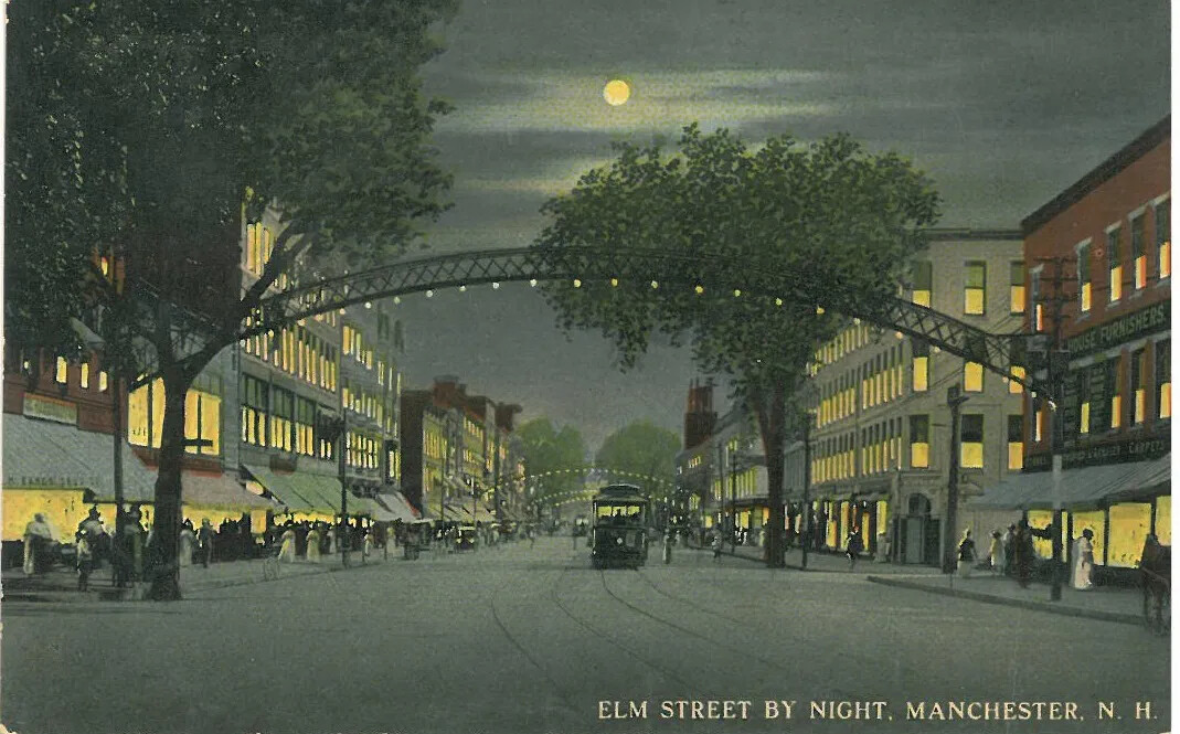 A colored postcard of a wide street lined with multi-storied buildings. The scene is depicted at night, with a full moon in a cloudy sky. All of the buildings have light shining through the windows. A trolley drives down the road with its headlights on. Several people walk along the sidewalks. Decorative iron arches with lightbulbs cross over the street at distanced intervals. The title of the postcard, located in the bottom right-hand corner, reads: "ELM STREET BY NIGHT, MANCHESTER, N. H."
