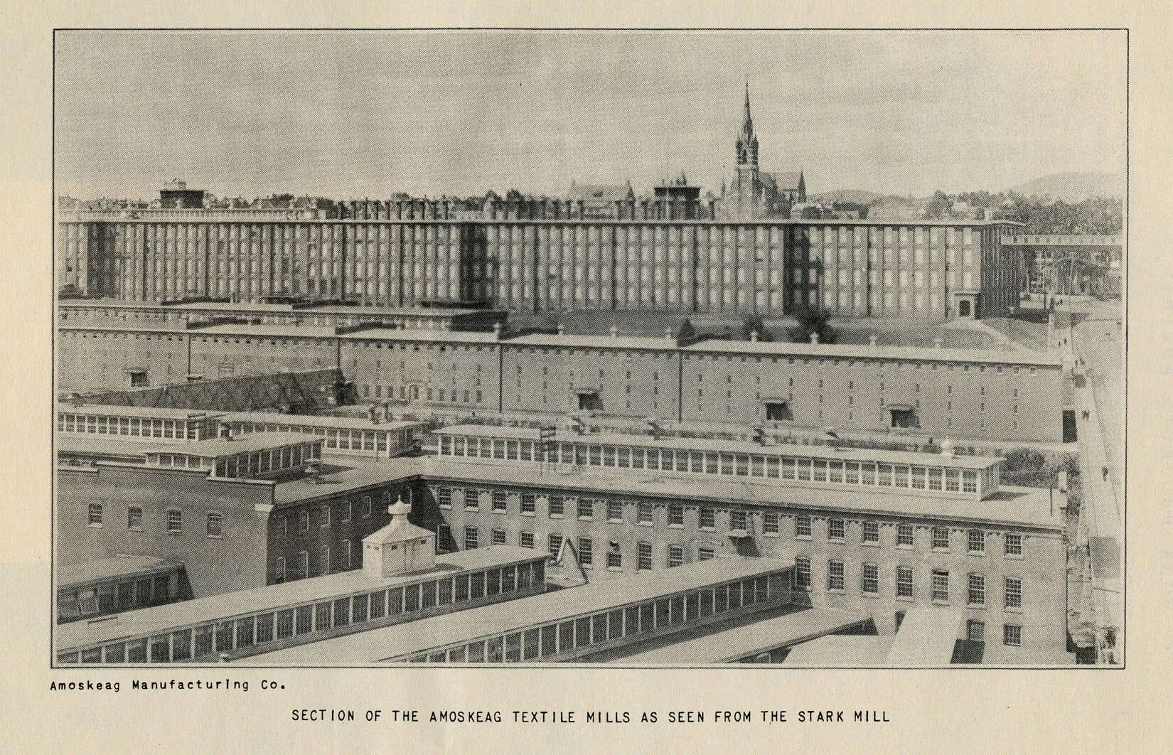 A black and white photograph shows many mill buildings from a high perspective. In the foreground, they run horizontally and vertically, and in the background there is one long factory building. Behind and on the sides of the buildings, the surrounding town can be seen. The caption reads "SECTION OF THE AMOSKEAG TEXTILE MILLS AS SEEN FROM THE STARK MILL" and on the left, "Amoskeag Manufacturing Co."   
