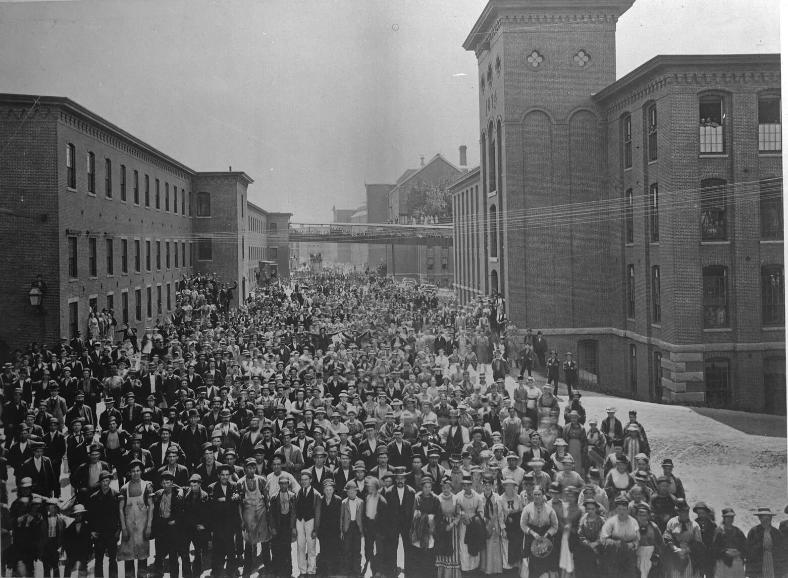 A black and white photograph of a large group of men, women, and children standing between two multi-storied buildings. Everyone in the photograph looks up at the camera. 