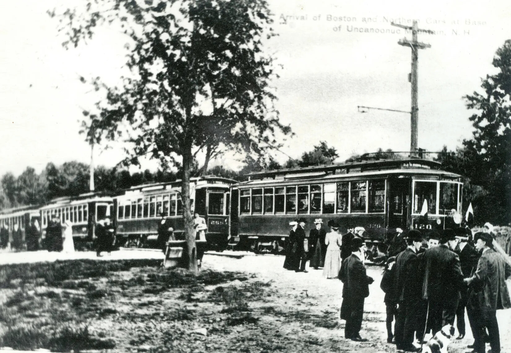 A black and white photograph of a group of people waiting in front of four trolleys. All of the men wear dark colored suits and bowler hats and the women wear long dresses and large, broad-rimmed hats.
