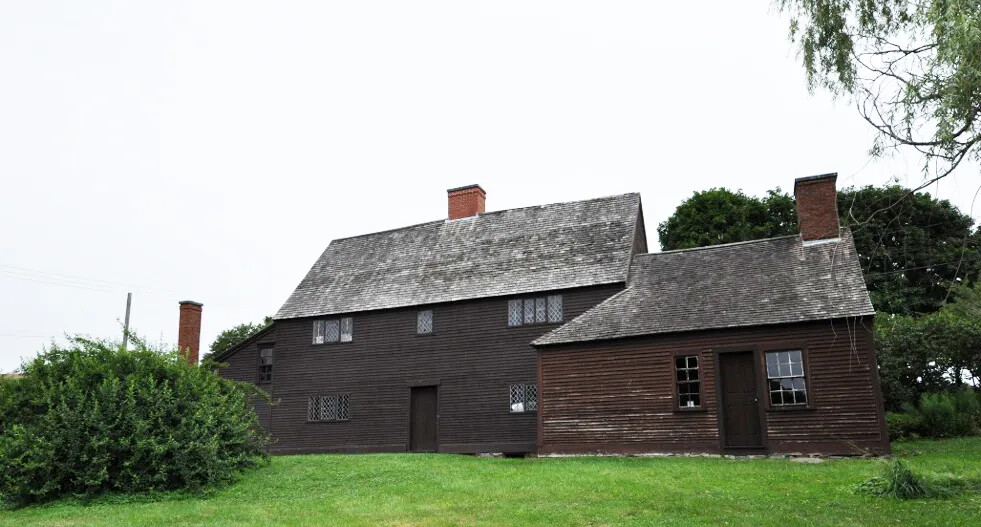 A colored photograph of a two-stored wooden house. The house sits atop a hill with green grass, bushes, and trees.
