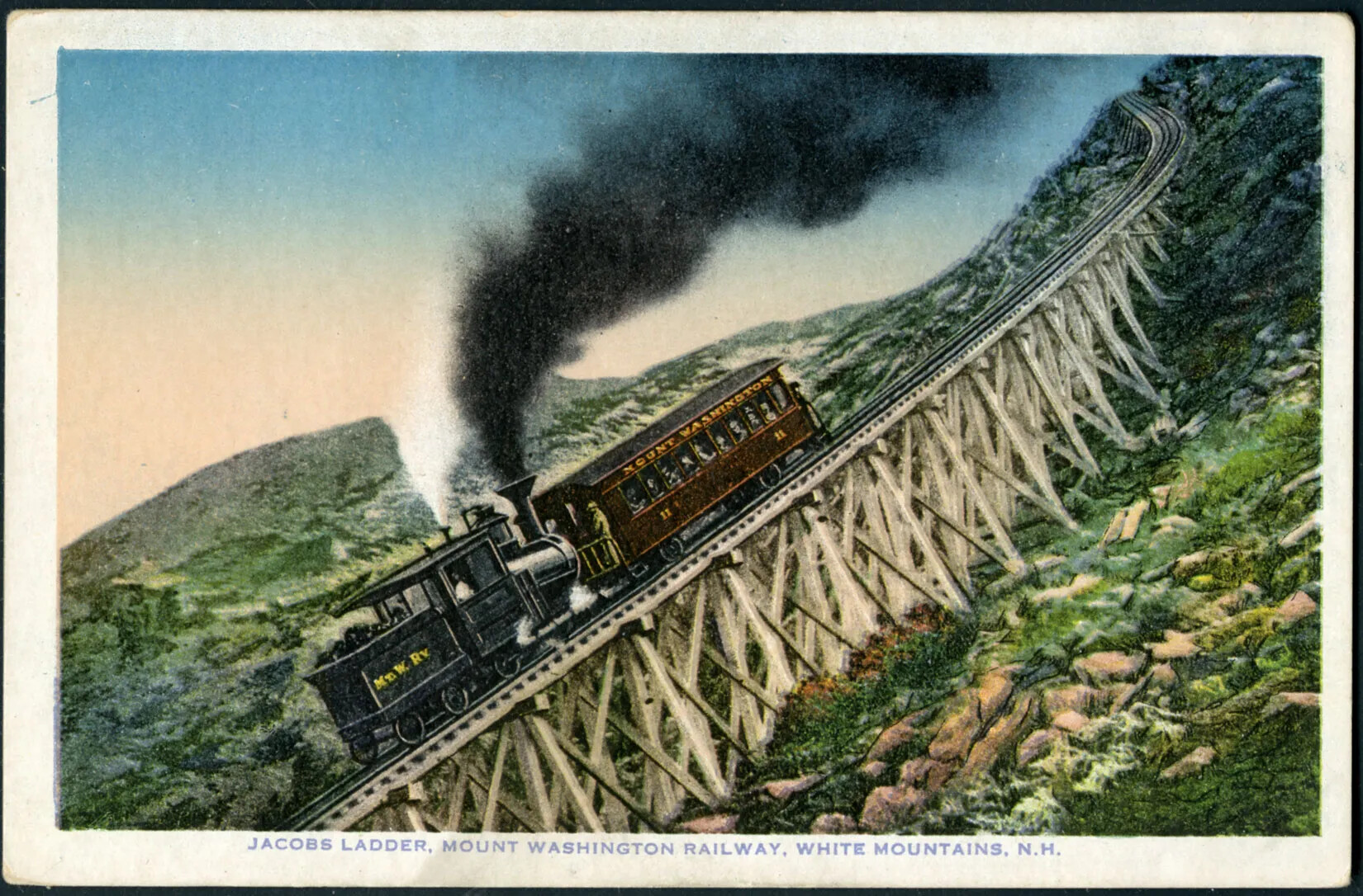 A color postcard of a train with one passenger car has a caption reading "Jacob's Ladder, Mount Washingon Railway, White Mountains, N.H." The train is descending from a very steep part of the tracks on the side of a mountain, which is covered with scrub and rocks. The part of the track the train is on is built up on a small bridge made of timber to keep the track as consistent as possible. 