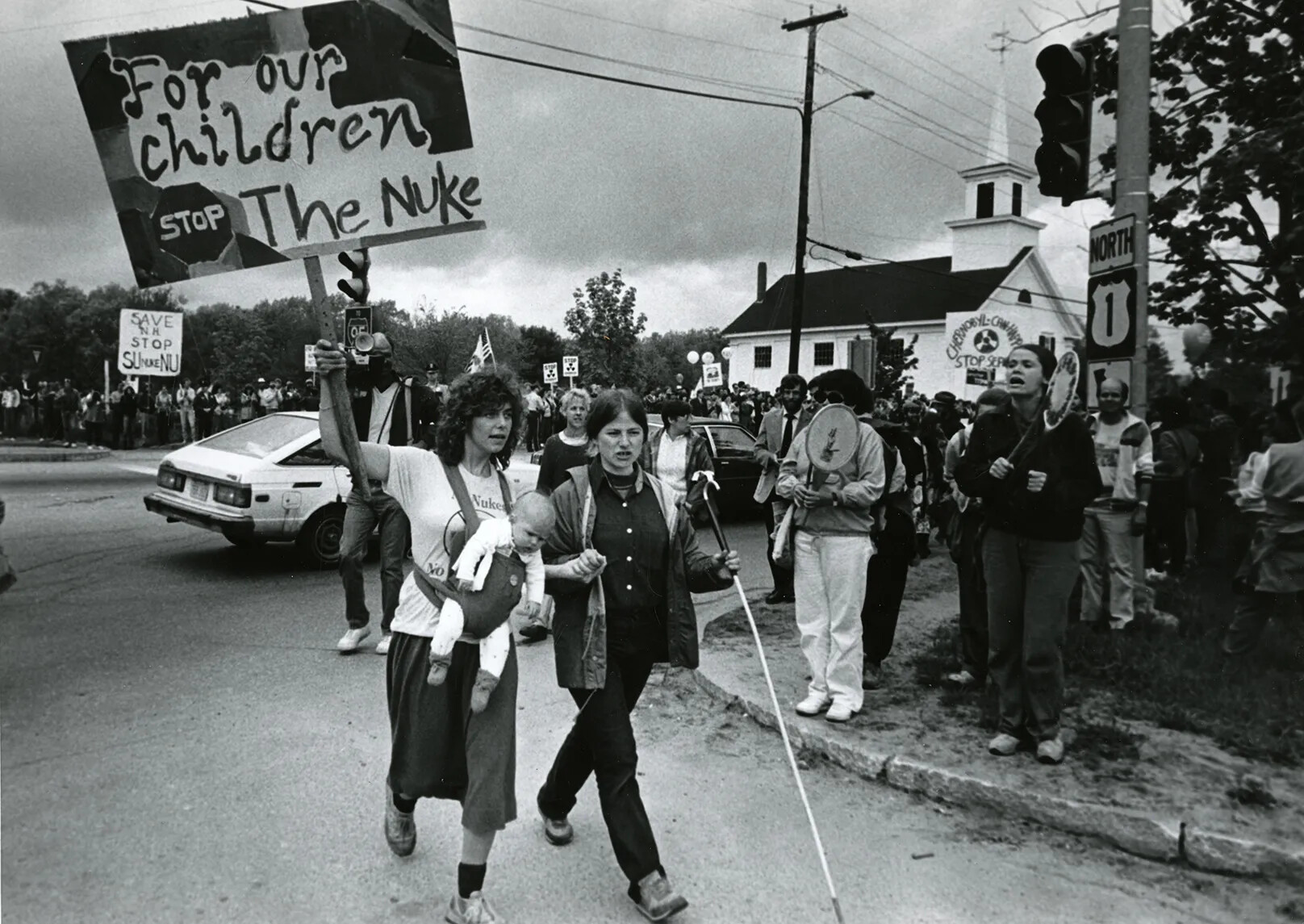 A black and white photograph of a crowd of people protesting on both sides of a paved road. Several people carry signs and look like they are saying something. A church with a tall steeple can be seen in the background.