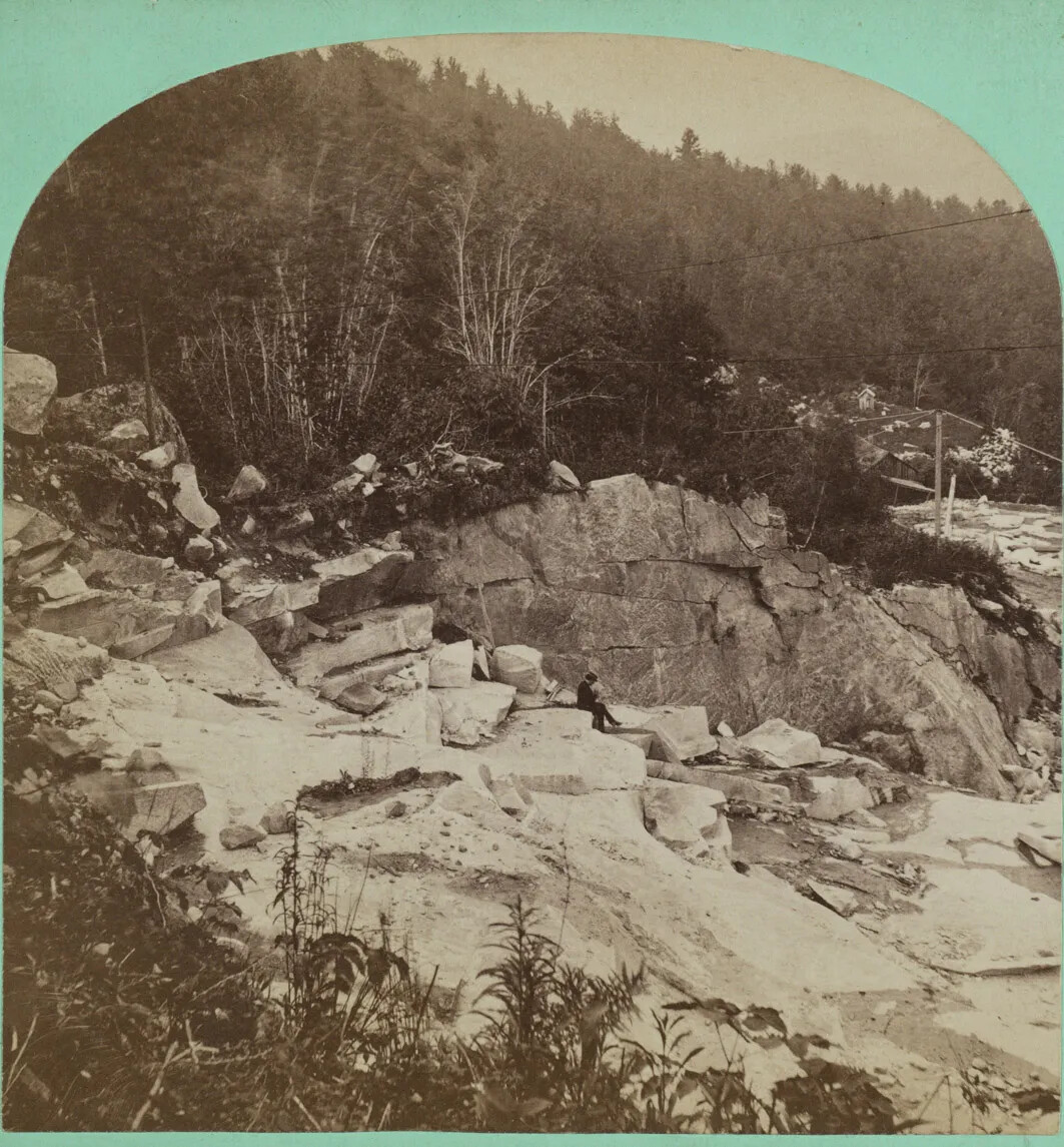 A square black and white image that is arched on top and bordered in teal on the top and bottom. The image shows the side of a hill, and the top and bottom of the image are wooded. In the middle is a wide swath of light colored rock. There are some large boulders, some slabs, and some cliff-like drops.