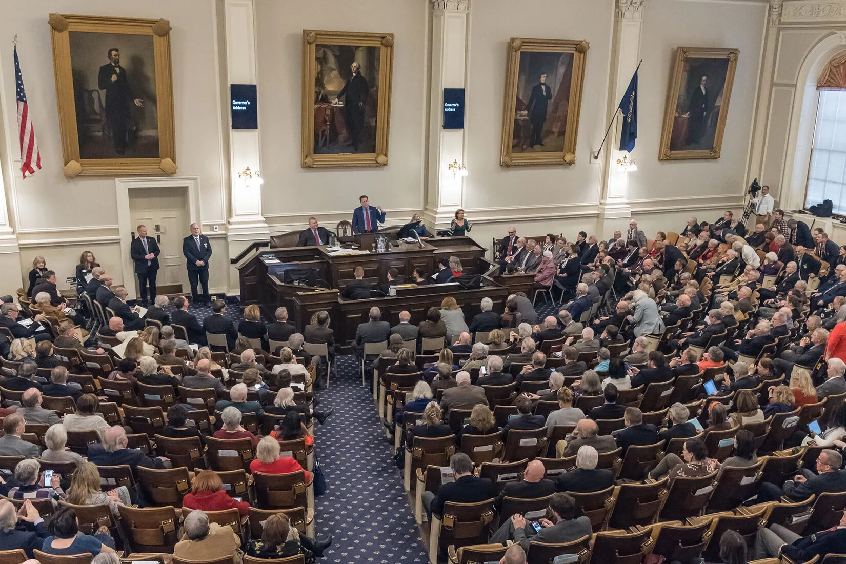 A New Hampshire governor addresses a session full of people. 