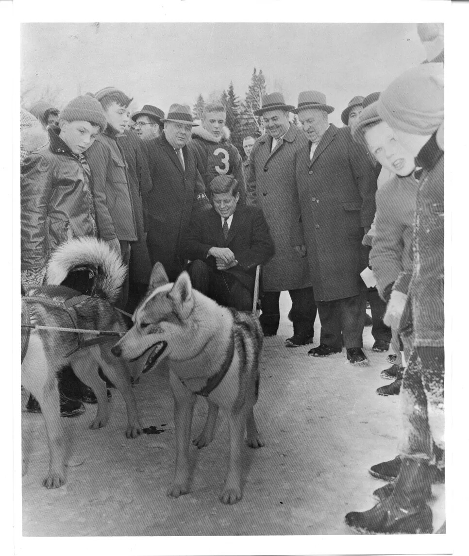 In a photograph, a smiling man in a suit is sitting in the sled while two sled dogs prepare to pull him. A crowd of men and boys in winter wear surround the man in the sled, watching.