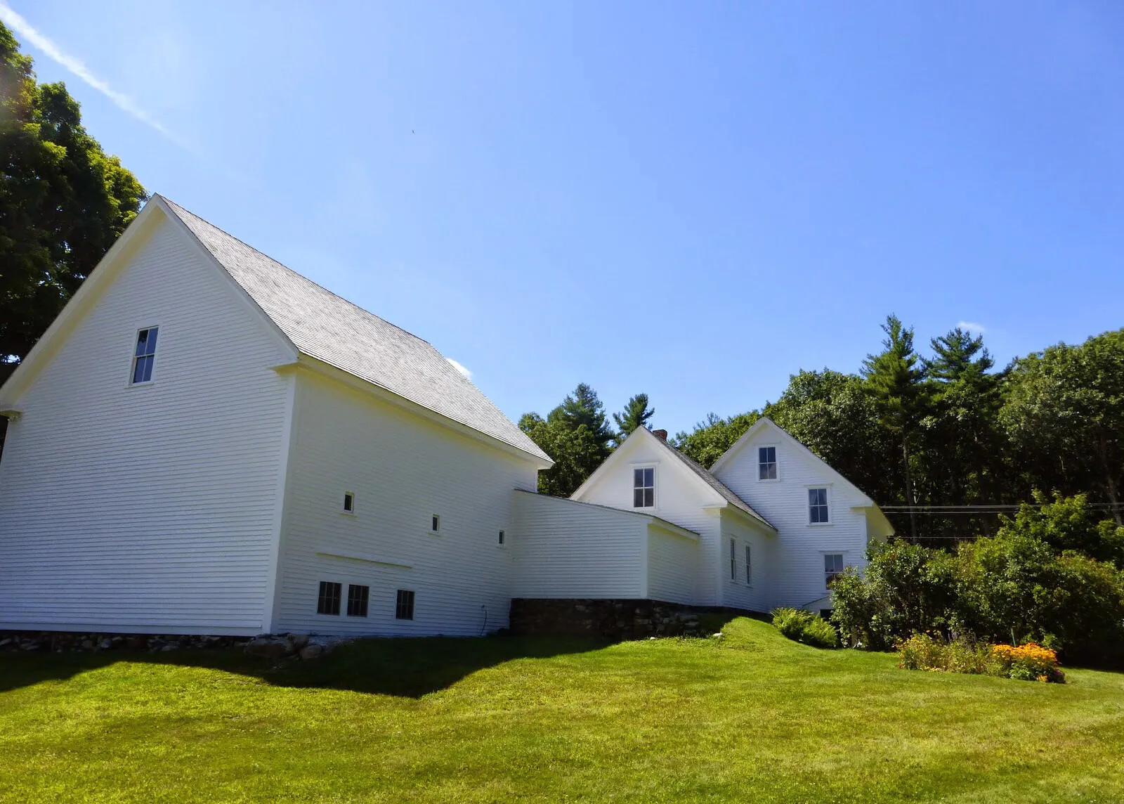 A colored photograph of a white two-storied house, with several multi-storied buildings attached to the back of it. Surrounding the house is a green field and tall trees with green leaves.