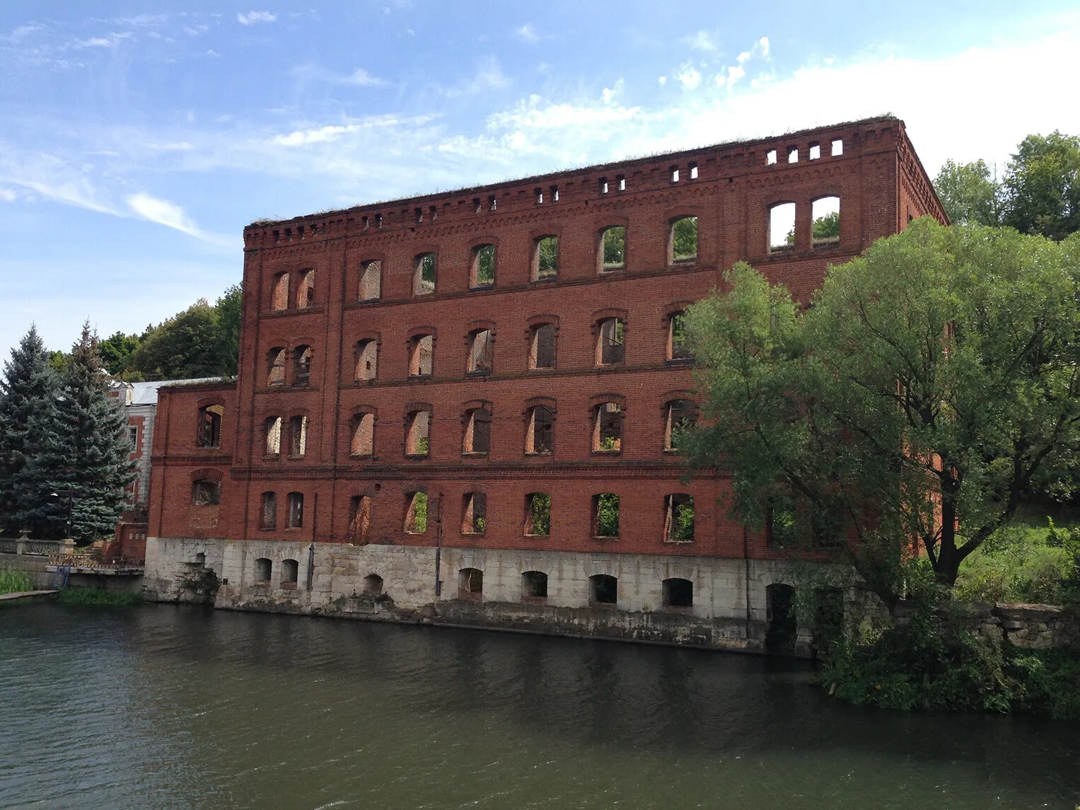 A colored photograph of a multi-storied brick building. The building no longer has a roof, and the woods behind the building can be seen through all of the windows. The building sits right on a body of water.