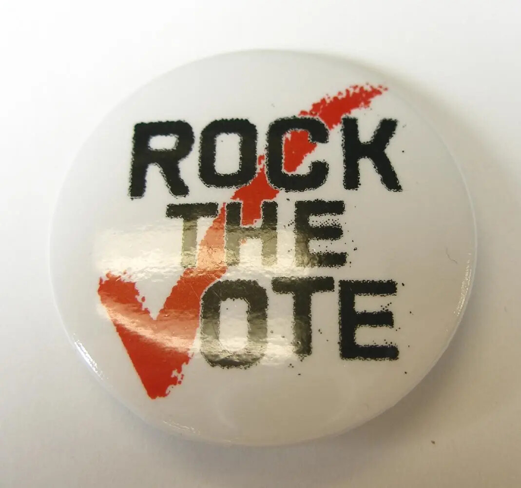 Circular metal Rock the Vote pin, black letters on white background with large red check mark forming the "V" in vote.
