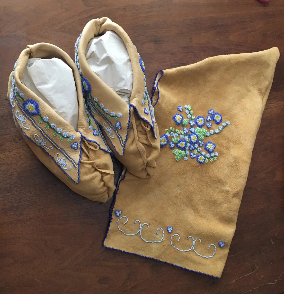 A pair of decorated brown leather moccasins and a small cap. The items are decorated with sewn designs of flowers, made from blue and green string.