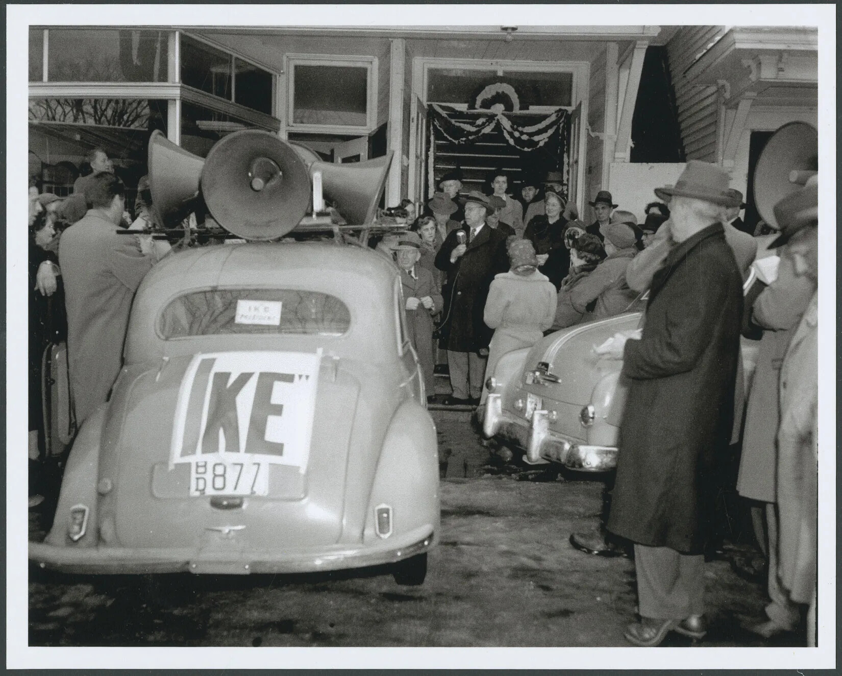 In a photograph of a street scene a man in an overcoat and hat stands near an open doorway on a sidewalk, holding a microphone and speaking to a crowd of people gathered around him. People wear raincoats, women are wearing hats or scarves over their heads, and men are wearing hats. A car parked at the curb has loudspeakers on its roof and a sign reading "Ike" taped to the trunk. 