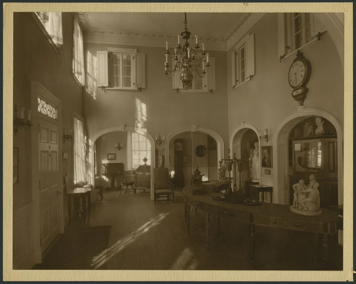 A black and white photograph of the entrance hallway of a two-storied building. The building's front door is on the left side of the photograph. A large semi-circular desk with various small statues and objects sits on the right-side of the photograph. Surrounding the room are several passage ways, which could be entered through arched doorways. The building's second floor, bordered by sets of windows and shutters, can be seen from this room.