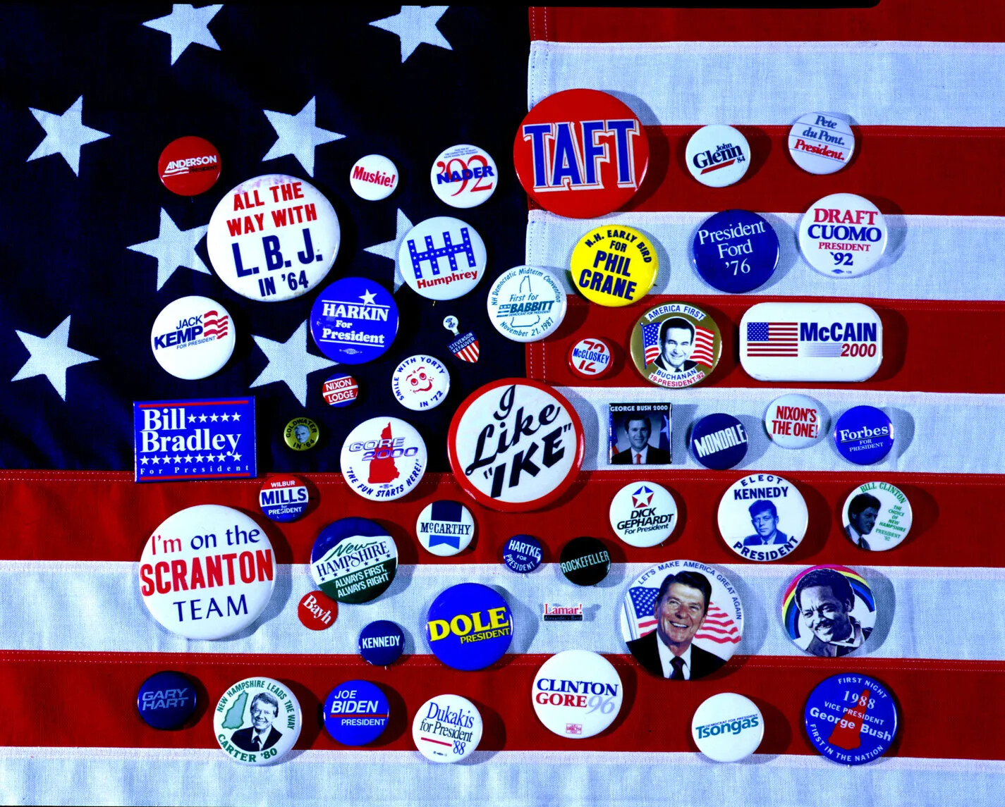 A photograph of a selection of more than 30 campaign buttons lie on part of an American flag. Buttons show faces, names, and slogans.