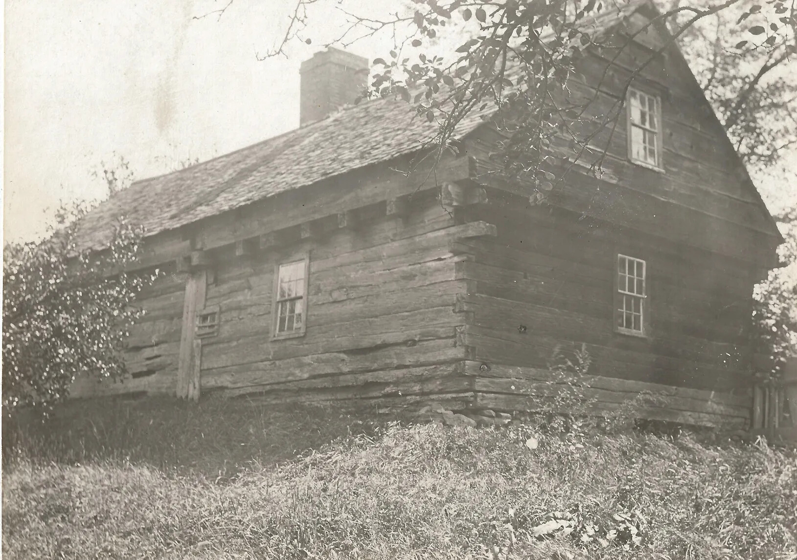A black and white photograph of a two-storied log cabin. The cabin sits in a field among tall bushes and trees.